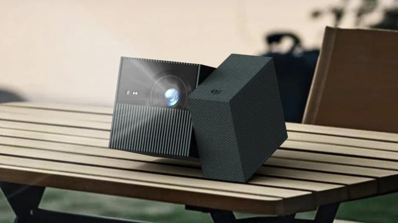 A TCL PlayCube sits outdoors on top of a table, and the image gives the appearance that the device is illuminating light from its projector