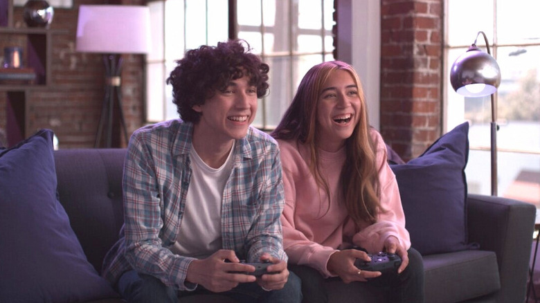 A teenage boy and girl sit on a sofa holding a game controller each