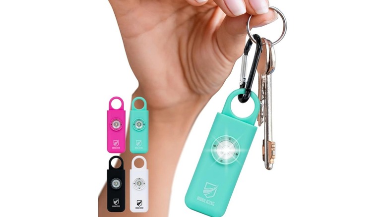 A woman holding a keychain to which a mint-colored Original Defense siren keychain is attached. The siren itself is made up of two parts, with a base body that has a light and an alarm and a hook that you can pull out to activate it