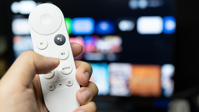 A person holding Google TV streamer remote with an out of focus TV in the background
