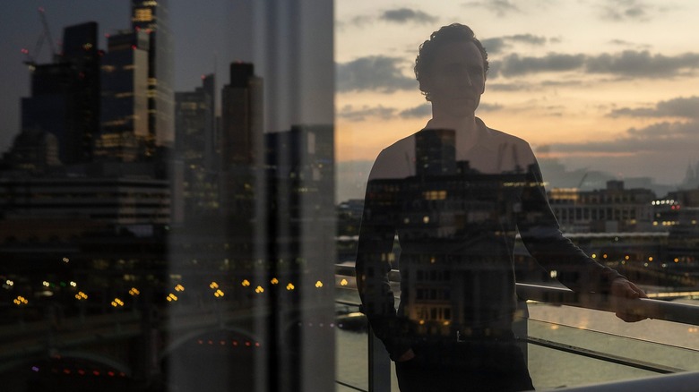 Tom Hiddleston stares contemplatively out onto a city scape at dusk.