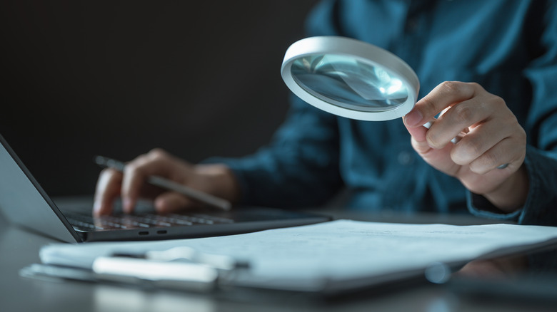 A person typing on his laptop, holding a magnifying glass in his other hand and looking at items on a clipboard