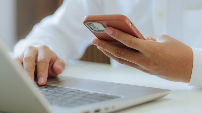 A person typing their computer while also looking at their phone