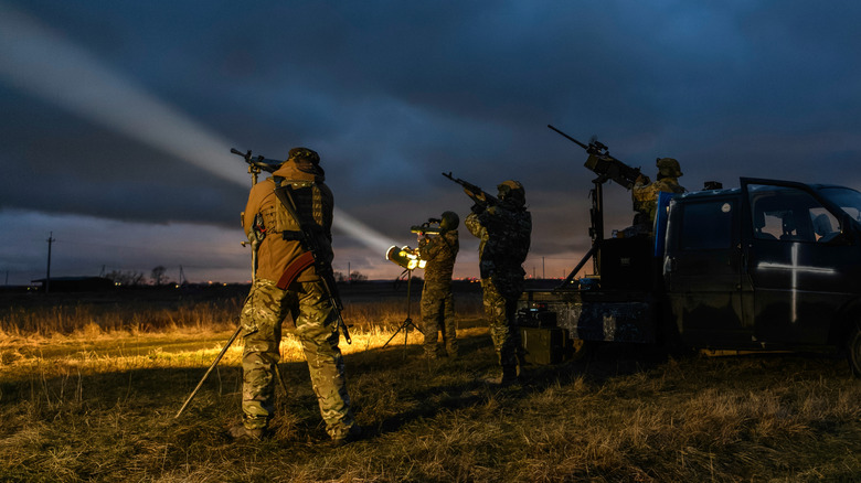 Ukrainian soldiers stand in a field and point search lights and artillery towards the night sky in search of killer drones.