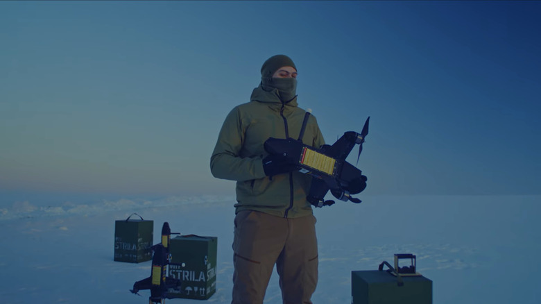 A man holds a Strilla anti-drone interceptor in an arctic landscape, where three boxes and two more drones sit behind him in the snow.