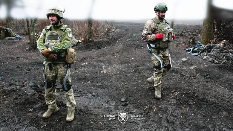 Two soldiers wearing exoskeletons around their legs walk through the battlefield.