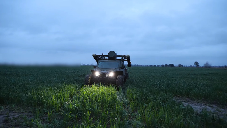 A buggy strapped with Hellfire missiles and radar atop its hood idles in a grassy field at dusk with its lights on.