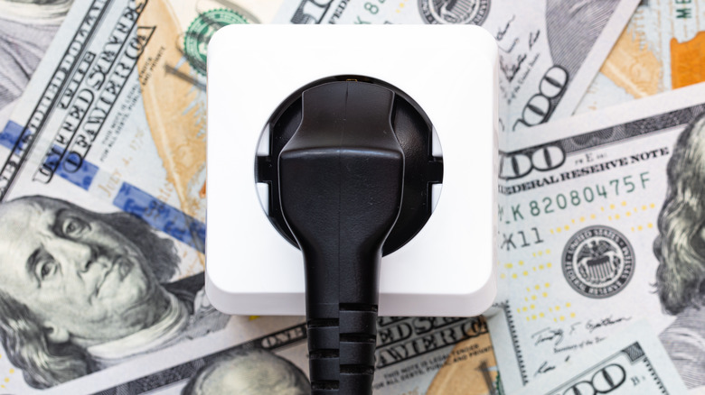 A pile of hundred dollar bills with a power cable attached to an outlet sitting on top of the money