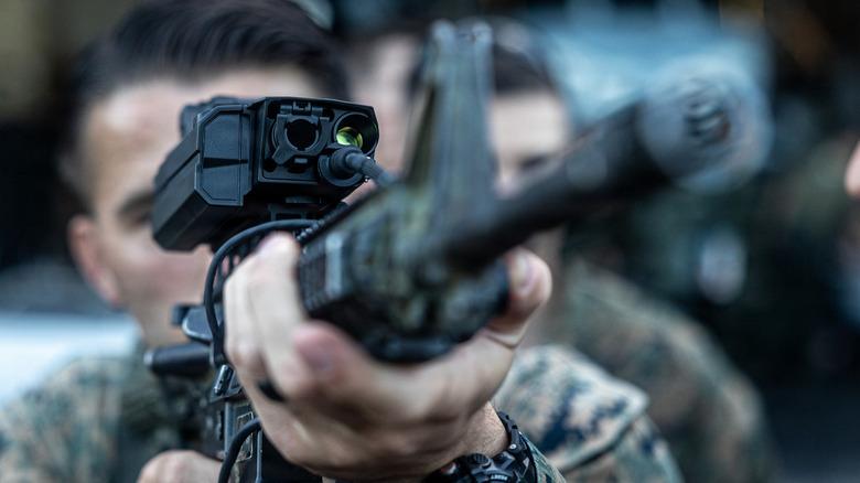 A dark haired marine in full camo points his rifle into the sky, showcasing the new black scope with sightlines and cameras.