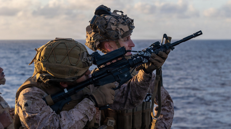 A marine aboard a naval ship points a rifle into the sky, looking through his smart scope, as another marine watches.