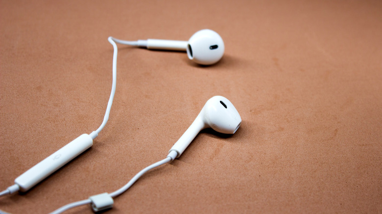 The image shows a pair of white wired earbuds lying on a brown surface. The earbuds have an in-line remote control on the cord, which is visible in the foreground.