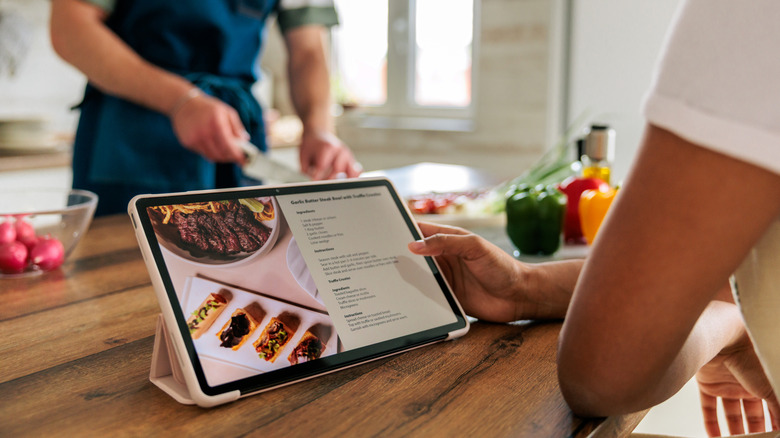 Hand holding kitchen display showing recipe with person preparing meal in the background