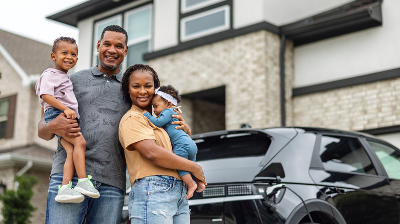 Smiling family with two children standing in front of their modern home and electric vehicle.