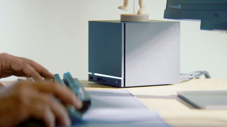A pair of hands resting on a mouse and keyboard with Valve's Steam Machine behind them visible in the background