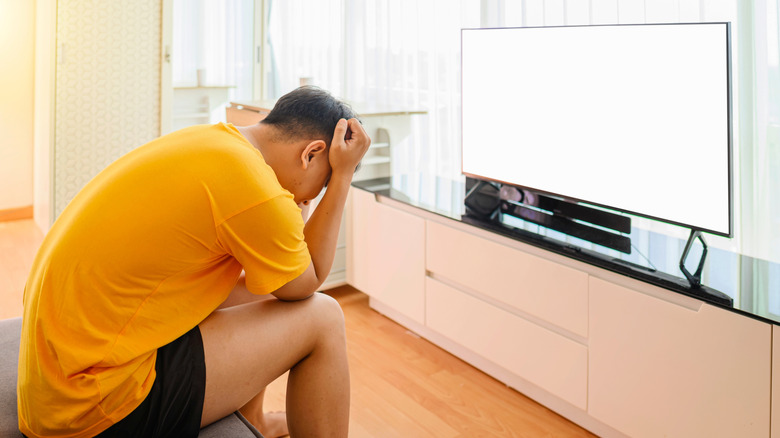 A frustrated man sits in front of a blank TV screen with his head between his fists.