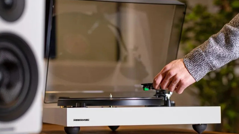 A Fluance turntable with a clear dust cover, shown as a hand lowers the tonearm onto a vinyl record