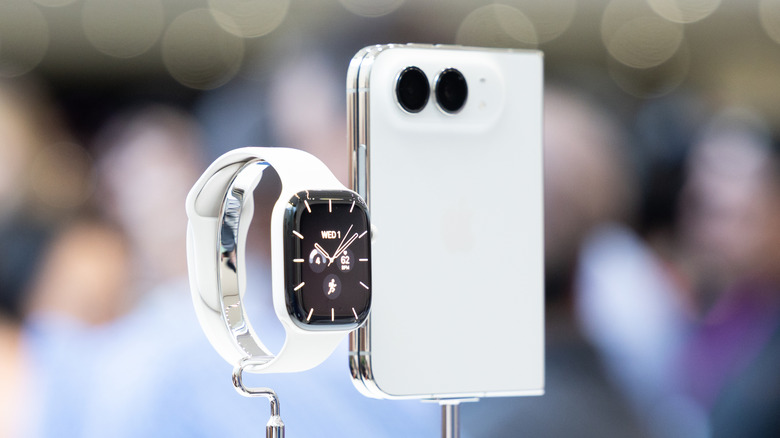 A Star White iPhone Duo sits atop a silver stand in its folded position, an Apple Watch 12 with a white band sits on its own stand next to it