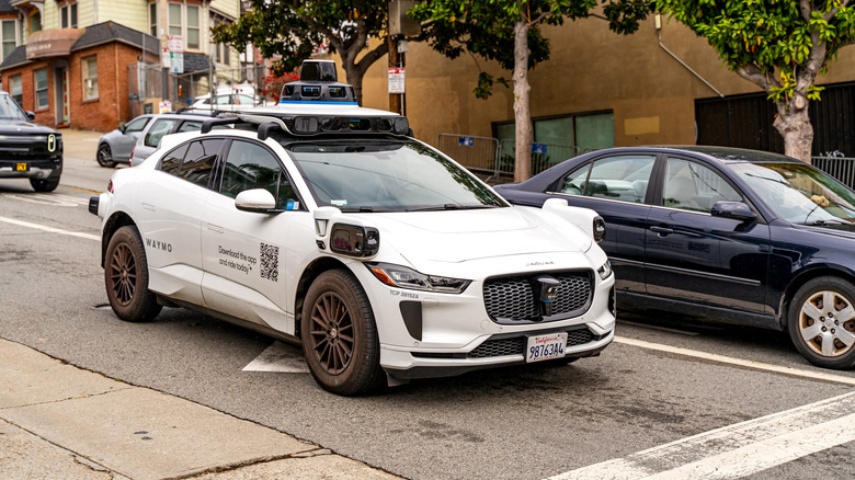 A driverless waymo taxi waits at an intersection next to a dark colored car
