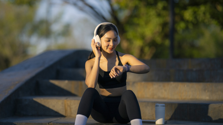 A woman in workout attire listening to music and looking at her smartwatch while sitting outdoors
