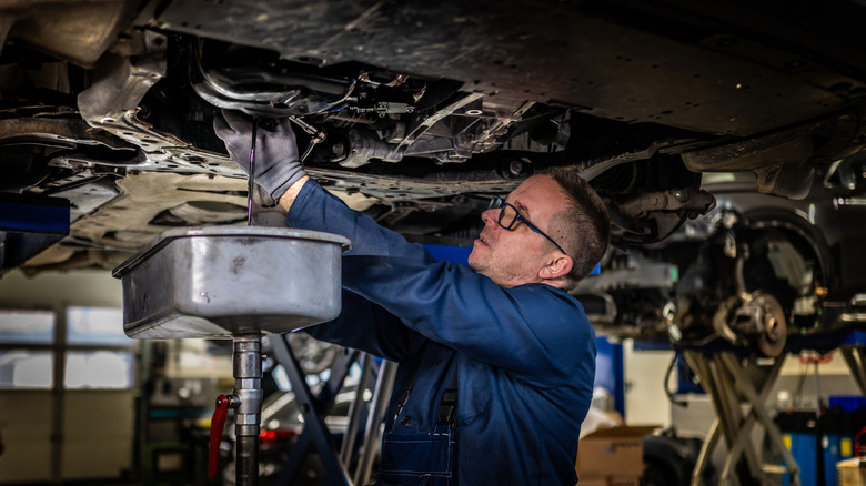 An auto mechanic changing the engine oil in a vehicle with a drip pan held below the drain.