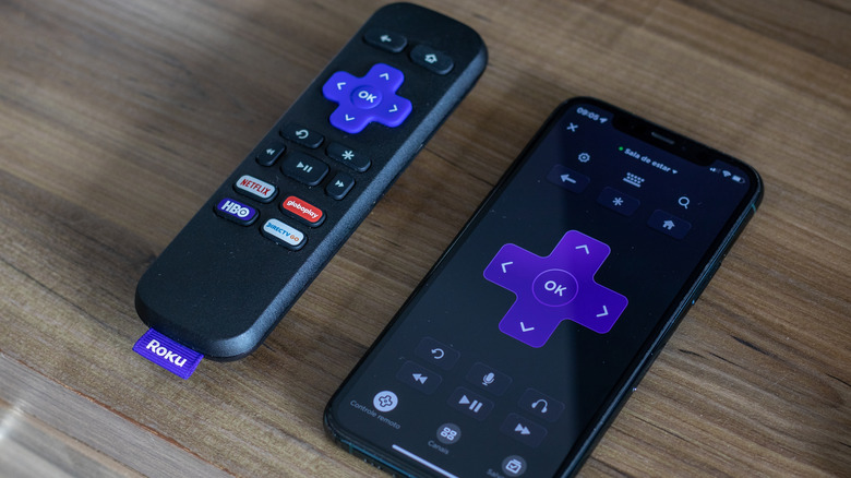A Roku remote placed next to a phone that has the Roku app opened.