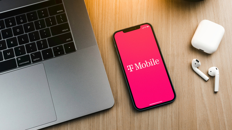 An iPhone with a wallpaper featuring T-Mobile's logo; between a Mac and a pair of AirPods.