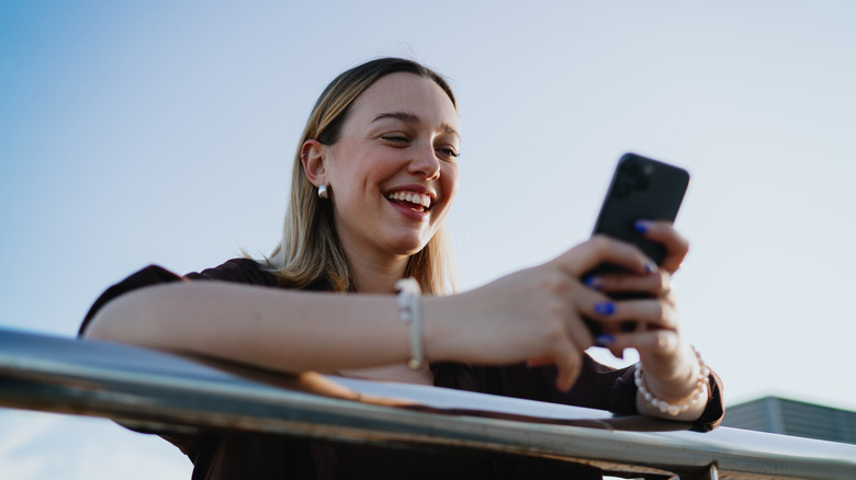 Joyful woman leaning on railing while browsing smartphone outdoors, enjoying digital world,