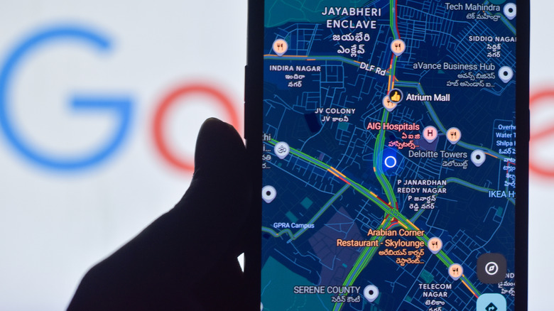 Closeup of a hand holding a smartphone showing Google Maps navigation display with a faint, blurred Google logo in the background