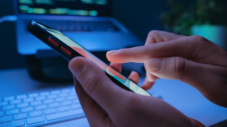 Hands holding a phone with a dark office setting, backlit by soft blue light, in the background