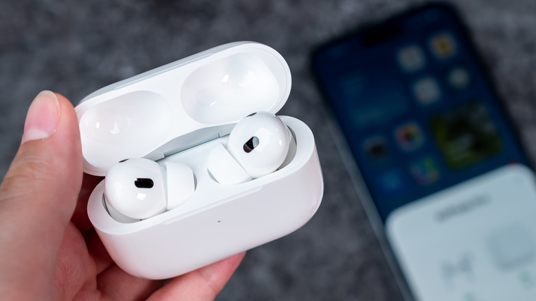 A person holding a pair of AirPods Pro in their charging case with the lid opened. An iPhone is placed on a surface beneath.
