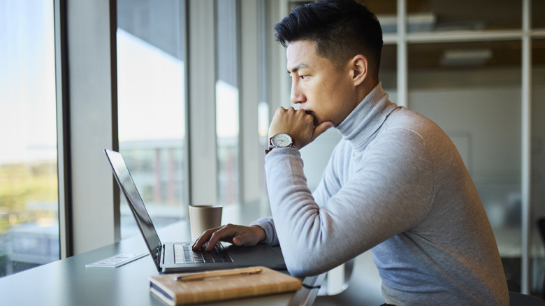 A man sits pensively looking at his laptop in front of a window.