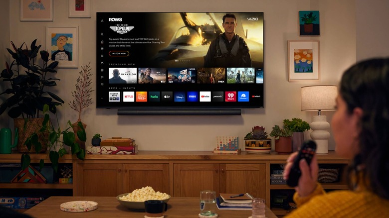 A wide-shot of a Vizio television displaying a variety of streaming content as an individual holds a TV remote as they sit in their living room filled with knick-knacks