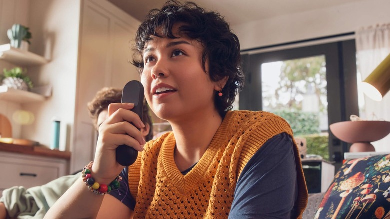 A closeup of an individual in a yellow sweater vest and blue T-shirt has a Vizio remote control in their hand and pointed straight to the ceiling