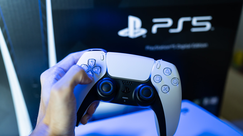 A  hand holding a DualSense controller with a blurred PS5 logo in the background