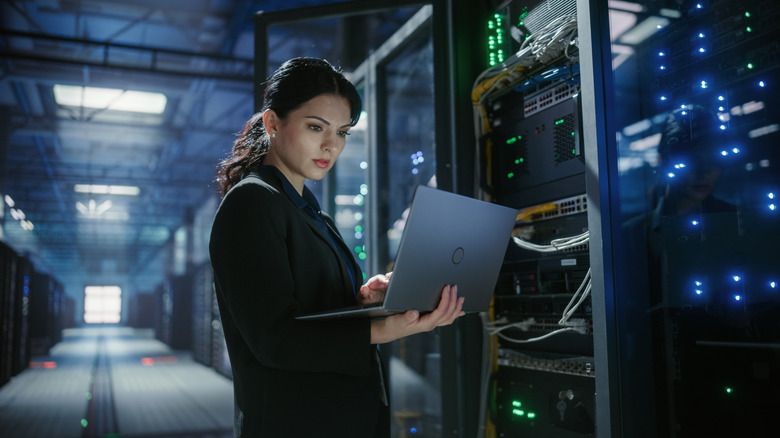 A woman holding a laptop standing in a server room.