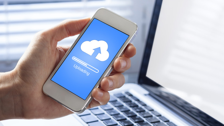 A hand holding a phone in front of a laptop, the phone screen showing uploading to the cloud.