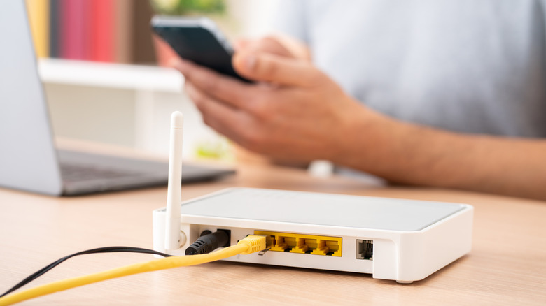 A close-up of a router on a desk with a person in the background using their phone near a laptop computer