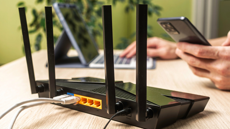 A close-up shot of a router on a desk with a person in the background using a phone and a computer