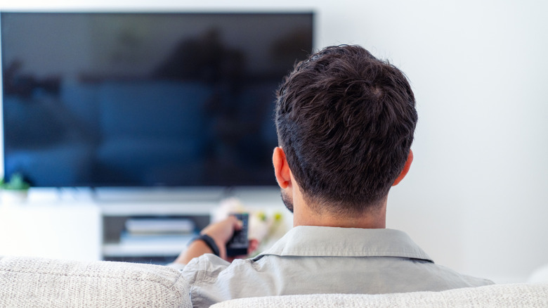 A man sitting on the couch, facing away from the camera, turning on a TV with a remote control
