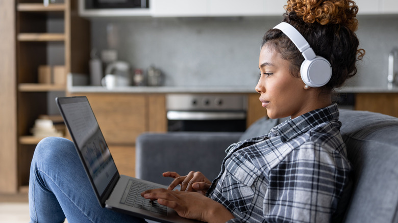 A young individual sits on a blue couch as they wear white headphones and navigate a laptop that rests on their lap