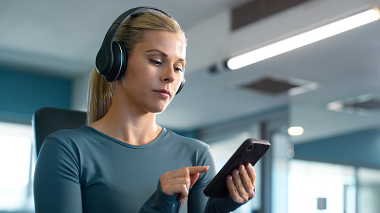 A woman wearing headphones and looking at a phone in her hand.