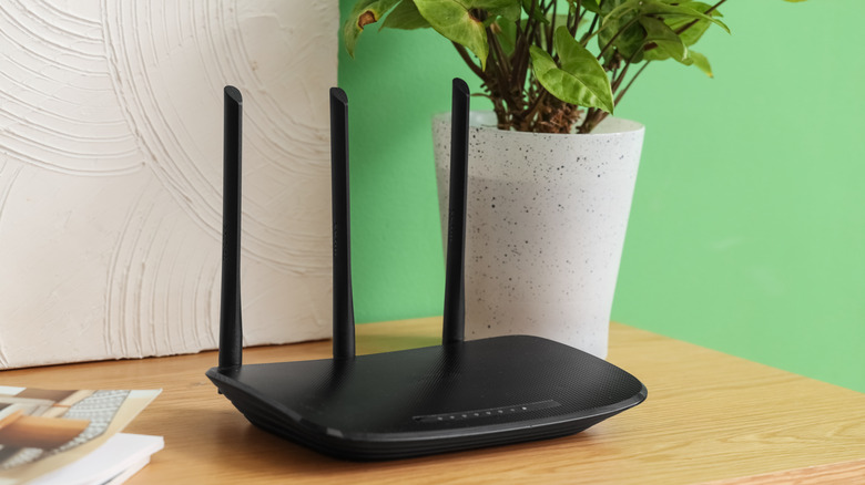 A black Wi-Fi router on a wooden table with a plant behind it.