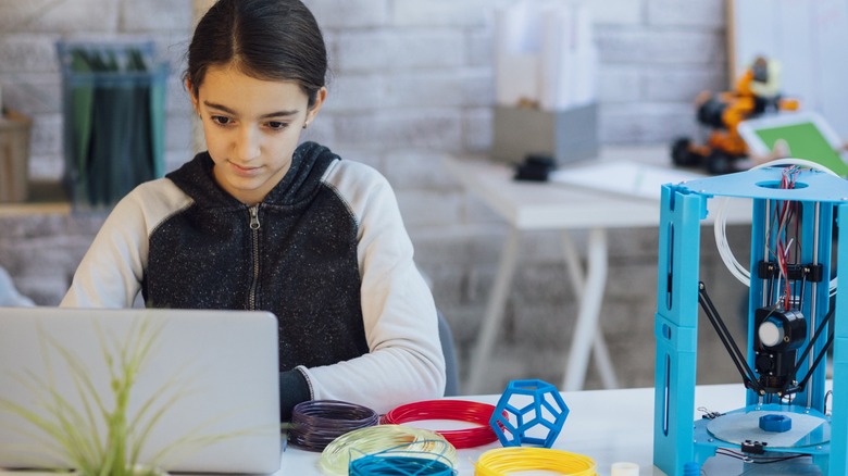 Photo of a girl sitting in her classroom and using laptop to programing 3D printer.