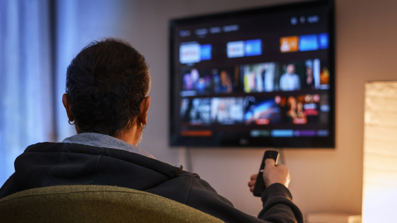 Man using an IR remote to select from streaming options on a wall-mounted smart TV