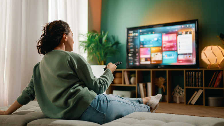 A woman sitting on a sofa and looking at her TV with a TV remote in her hand