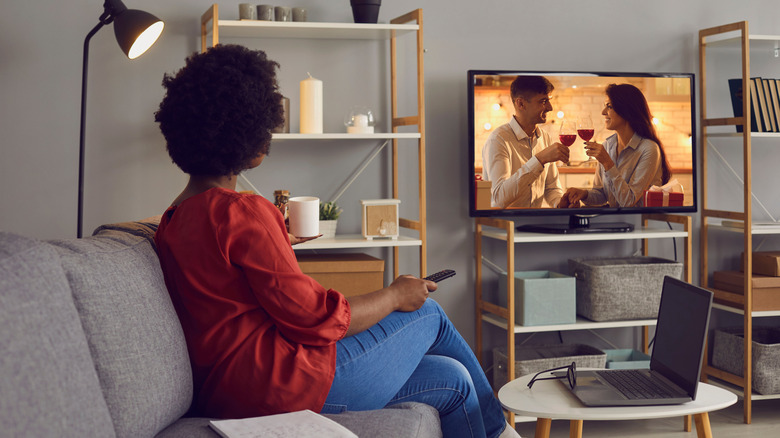 A woman sitting on the couch holding a remote and watching a soap opera on TV.