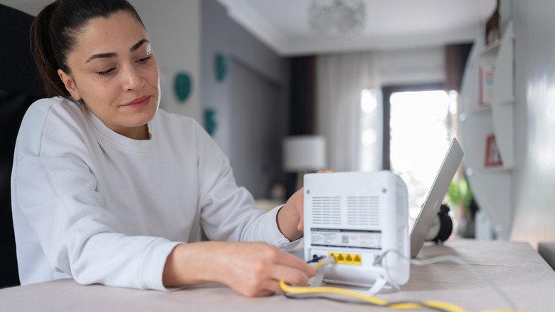 A woman plugging Ethernet ports into a white Wi-Fi router next to a computer.