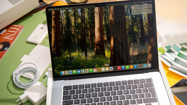 A MacBook displays its desktop screen while it is surrounded by by an assortment of items, including a charger and its adapter, a white iPhone, and more