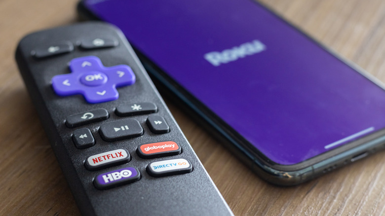 A Roku remote placed next to a smartphone loading the Roku mobile app, both sitting on a wooden table.