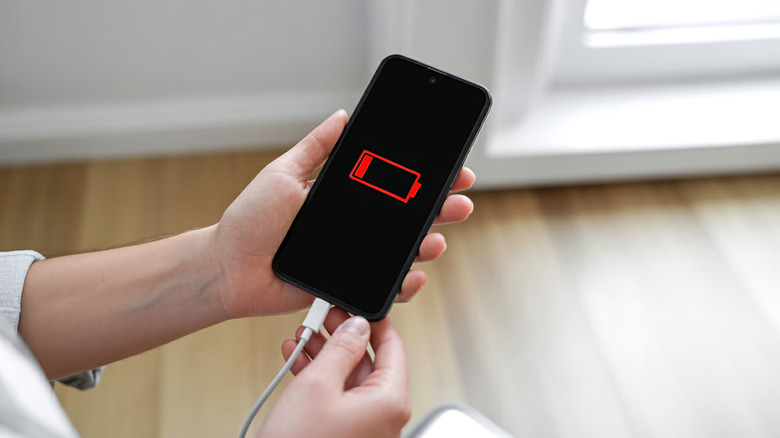 A person holding a phone plugged into a charger with a low battery onscreen indicator.